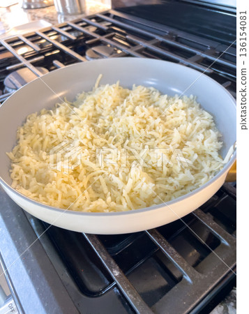 A pan on a stovetop is filled with shredded white rice being cooked. The scene is brightly lit and shows the rice spread evenly inside the pan. The stove surface and kitchen setting are clearly A pan on a stovetop is filled with shredded white rice being cooked. The scene is brightly lit and shows the rice spread evenly inside the pan. The stove surface and kitchen setting are clearly 136154081