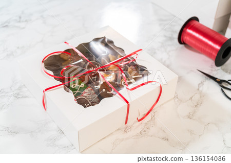 Captured from a close-up perspective, white holiday cookie boxes with elegant cut-out windows reveal intricately decorated Christmas cookies inside. The red ribbon adds a festive touch, making these Captured from a close-up perspective, white holiday cookie boxes with elegant cut-out windows reveal intricately decorated Christmas cookies inside. The red ribbon adds a festive touch, making these 136154086
