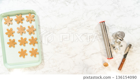 Overhead view of amber gingerbread dough shaped into snowflake cookies arranged neatly on a green baking tray, with rolling pins and cutters placed nearby. 136154090