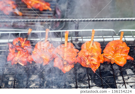 Authentic Thai grilled chicken (Kai Yang) skewers cooking on a charcoal grill with smoke. Delicious traditional Thai street food and local culinary culture 136154224