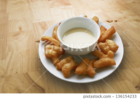 Deep-fried dough sticks or Pa Tong Go served with sweetened condensed milk on a white plate. Traditional Asian breakfast on a wooden table. Delicious Thai street food snack Deep-fried dough sticks or Pa Tong Go served with sweetened condensed milk on a white plate. Traditional Asian breakfast on a wooden table. Delicious Thai street food snack 136154233