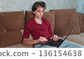 A young man is focused on his laptop while seated comfortably on a brown couch. guy is working on a laptop. A teenage boy is studying remotely at home using a computer. 136154638