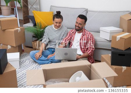 Home Happiness: A cheerful young couple, amid moving boxes in their new apartment, uses a laptop to shop for decor, create their dream space, or plan their move. 136155335