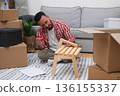 Self-Made Home: Young man constructs a DIY shelf in his new residence, surrounded by moving carton boxes, embracing the journey of relocating 136155337