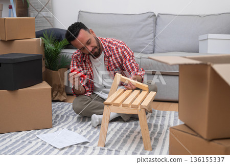 Self-Made Home: Young man constructs a DIY shelf in his new residence, surrounded by moving carton boxes, embracing the journey of relocating 136155337