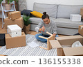 woman, amid moving carton boxes and furniture, sits on the floor to assemble a self-assembly shelf in her rental house 136155338