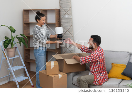 delighted young family, husband and wife, enjoys the process of moving into their new residence, placing a lamp on an empty shelf and embracing the joy of relocation delighted young family, husband and wife, enjoys the process of moving into their new residence, placing a lamp on an empty shelf and embracing the joy of relocation 136155340