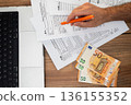 Top view of a person filling out tax forms with an orange pen, euros, and a laptop on a wooden desk. Financial accounting and tax declaration process  136155352