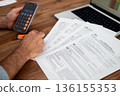 Man working on tax documents with calculator and laptop on wooden table. Financial analysis and tax preparation concept  136155353