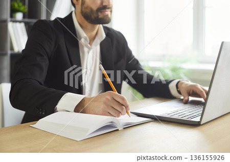 Focused businessman writing notes at desk with laptop in bright office workspace Focused businessman writing notes at desk with laptop in bright office workspace 136155926