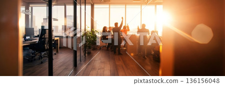 Group of businesspeople celebrating success with arms raised in a modern office with large windows and sunset view, showing teamwork, achievement, and positive work environment 136156048