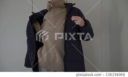 Woman putting on winter jacket over beige turtleneck sweater indoors Woman putting on winter jacket over beige turtleneck sweater indoors 136156068