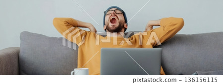 Tired man wearing glasses and a beanie yawns while sitting on a couch with a laptop, indicating stress and fatigue from multitasking and managing deadlines in a remote work environment 136156115