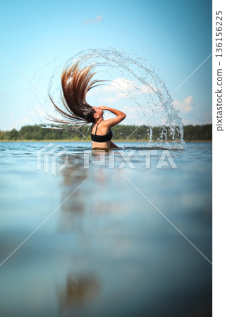 Girl splashing water with hair flip in lake, summer joy and freedom. Vertical summer background. Girl tossing wet hair in lake splash, sunny summer vibe Girl splashing water with hair flip in lake, summer joy and freedom. Vertical summer background. Girl tossing wet hair in lake splash, sunny summer vibe 136156225