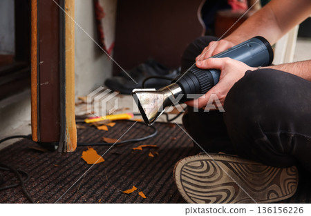 Removing Old Paint with Heat Gun. A close-up of a person using a professional electric heat gun to strip old, peeling brown paint from a wooden door frame. a home renovation or restoration project Removing Old Paint with Heat Gun. A close-up of a person using a professional electric heat gun to strip old, peeling brown paint from a wooden door frame. a home renovation or restoration project 136156226