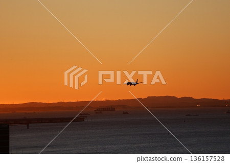 Orange sunrise sky and a silhouette of an airplane preparing to land, mountains, the sea, and a ship 136157528