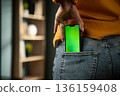 Unrecognizable woman slipping a modern smartphone with green screen into her jeans back pocket, close-up lifestyle shot ideal for mockups, tech, app and advertising concepts 136159408