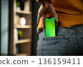 Hand sliding a modern smartphone with a vivid green screen into a rear jeans pocket, symbolizing privacy, convenience, and technology integration in daily life 136159411