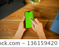 Woman's hands holding a modern smartphone with vivid green chroma key screen, tapping mockup over wooden desk with laptop and plant, top-view workspace for app or ad mockup 136159420