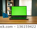 Laptop with a bright green screen for chroma key compositing sitting on a wooden desk in a modern home office with a bookshelf and window blinds in the background 136159422