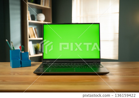 Laptop with a bright green screen for chroma key compositing sitting on a wooden desk in a modern home office with a bookshelf and window blinds in the background 136159422