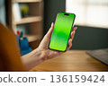 Person's hand holding a modern smartphone with a vivid green screen display, allowing for mockup customization in a home or office environment, representing modern technology and communication 136159424