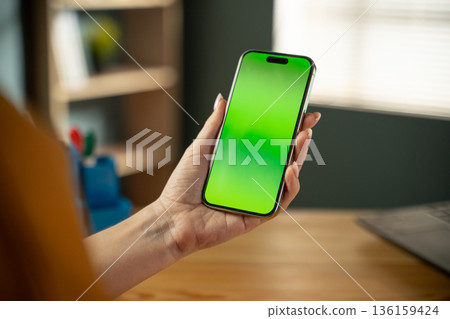 Person's hand holding a modern smartphone with a vivid green screen display, allowing for mockup customization in a home or office environment, representing modern technology and communication 136159424