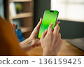 Woman's hands are holding a modern smartphone with a blank green screen. Actively tapping and interacting with the display for a mobile application or digital content. With a blurred office background 136159425