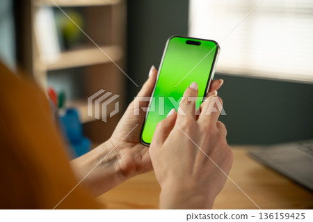 Woman's hands are holding a modern smartphone with a blank green screen. Actively tapping and interacting with the display for a mobile application or digital content. With a blurred office background 136159425