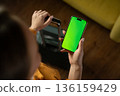 Young adult woman holding a modern smartphone with a blank green screen while pulling a suitcase, preparing for departure or planning a trip using a mobile application 136159429