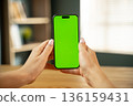 Woman holding a modern smartphone with a vivid green screen display, perfect for showcasing content, applications, or websites with a customizable chroma key background for creative projects 136159431