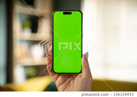 Adult hand holding a vertical modern smartphone with a vivid green screen for easy content replacement, simulating an immersive experience in a home environment Adult hand holding a vertical modern smartphone with a vivid green screen for easy content replacement, simulating an immersive experience in a home environment 136159433