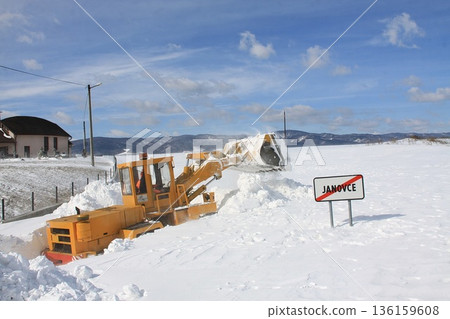 A yellow snowplow is clearing a village road after a snowstorm, which is completely covered with a mountain of snow. 136159608