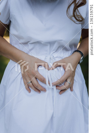 Pregnant woman in white dress holding belly in heart shape, maternity closeup, copy space 136159791