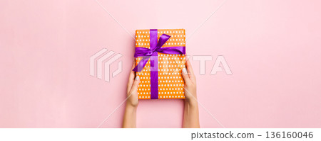 Female's hands holding striped gift box with colored ribbon on living coral background. Christmas concept or other holiday handmade present box, concept top view with copy space 136160046
