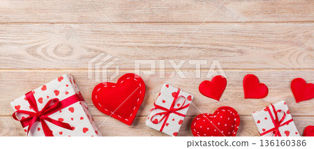 Valentine or other holiday handmade present in paper with red hearts and gifts box in holiday wrapper. Present box of gift on orange wooden table top view with copy space, empty space for design Valentine or other holiday handmade present in paper with red hearts and gifts box in holiday wrapper. Present box of gift on orange wooden table top view with copy space, empty space for design 136160386