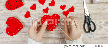 Valentine day theme. Workplace for preparing handmade decorations. Top view of female hands sew felt heart. Packed gifts, tools shabby wooden table Valentine day theme. Workplace for preparing handmade decorations. Top view of female hands sew felt heart. Packed gifts, tools shabby wooden table 136160429