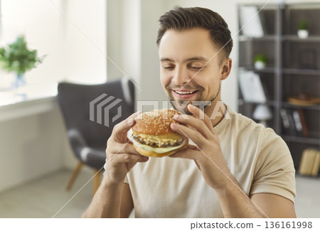 Happy hungry man sitting at home, office holding large tasty burger looking smiling happily Happy hungry man sitting at home, office holding large tasty burger looking smiling happily 136161998
