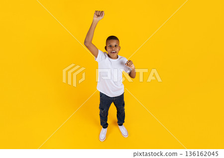 Black preteen boy raising fist in celebration, standing on yellow studio background 136162045