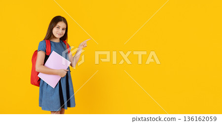 Preteen girl pointing aside with school backpack on yellow studio background 136162059