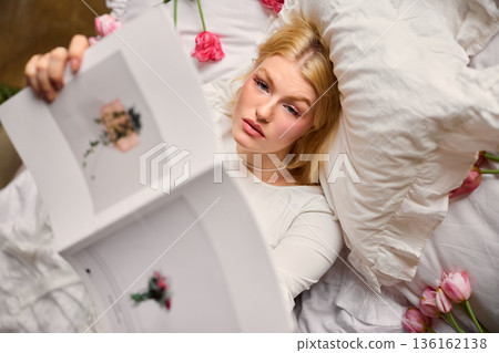 Serene woman reclining on white bedding surrounded by colorful tulips with thoughtful expression Serene woman reclining on white bedding surrounded by colorful tulips with thoughtful expression 136162138