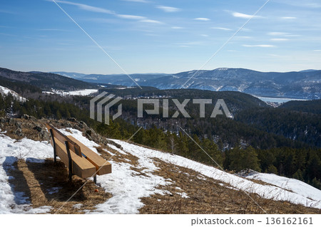 Empty bench on a snowy mountain with forest and town in background. Winter landscape for travel and nature exploration concept. 136162161