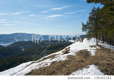 Empty wooden bench on a snowy hill overlooking a winter forest valley. Beautiful landscape for travel and nature exploration. 136162163