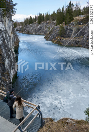 Man and woman enjoying view of partially frozen lake surrounded by rocky cliffs and pine forest in Ruskeala Park, cold weather tourism, travel destination concept 136162175