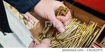 A man's hand picking up a handful of metal nails from a cardboard box at a hardware store or warehouse. Concept art of handicrafts, building materials, craftsmanship, and industrial supplies. 136162475