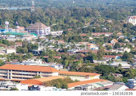 High-angle view of a provincial town in Thailand featuring residential houses, schools, and public buildings surrounded by lush green trees and tropical vegetation under bright sunlight. High-angle view of a provincial town in Thailand featuring residential houses, schools, and public buildings surrounded by lush green trees and tropical vegetation under bright sunlight. 136162636