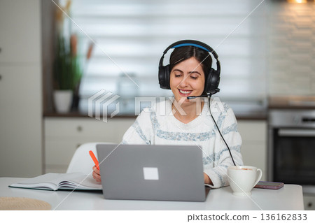 Young adult woman working remotely from home with headset and laptop Young adult woman working remotely from home with headset and laptop 136162833