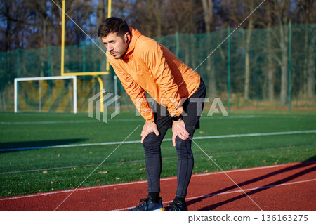 Man in orange sports jacket leaning forward with hands on knees on outdoor running track 136163275