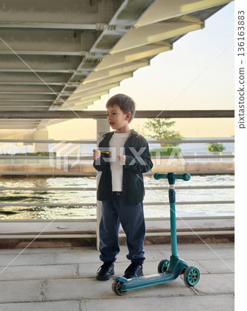 Young Boy Engaged In Bubble-Blowing While Standing By His Scooter. 136163883