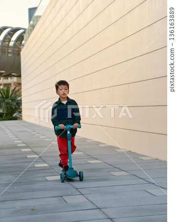 Young Child Riding A Scooter Down A Sleek, Modern Walkway. 136163889